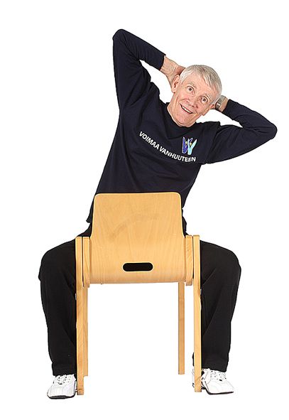 Jumppaaja istuu ryhdikkäästi tuolilla kasvot selkänojaan päin ja taivuttaa ylävartaloa vasemmalle sivusuuntaan.