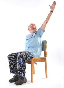 Jumppaaja istuu ryhdikkäästi tuolissa ja kurkottaa vasemmalla kädellä takaviistoon selän taakse peukalon osoittaessa kohti kattoa