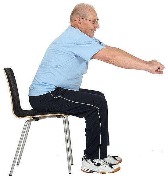 Jumppaaja istuu selkä irti tuolin selkänojasta. Hän nojaa ylävartalolla eteenpäin ja on ojentanut kädet suoriksi eteen vaakatasoon pitäen käsiä nyrkissä.