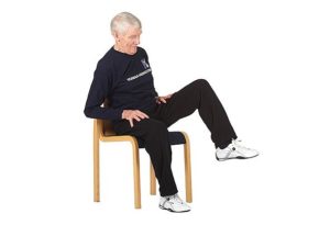 Jumppaaja istuu tuolilla ja on viemässä vasenta polvea sivulle painon ollessa oikealla kankulla.