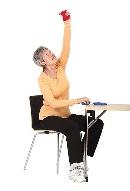 Jumppaaja istuu tuolilla ottaen oikealla kädellä tukea edessä olevasta pöydästä ja on ojentanut vasemman käden suoraksi ylös