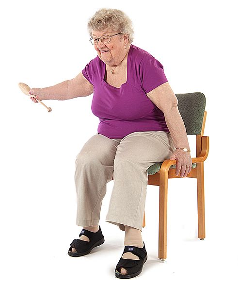 Jumppaaja istuu tuolilla ja on ojentanut oikean käden vasemmalle sivulle vaakatasoon keila kädessään ja pitää vasemmalla kädellä kiinni tuolin reunasta.