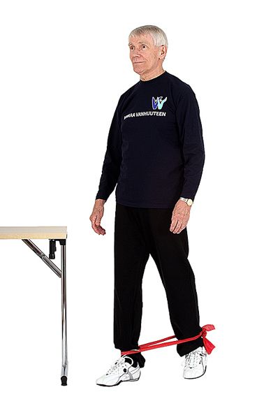 Jumppaaja seisoo ryhdikkäästi kuminauha nilkkojen ympärillä ja on astunut vasemmalla jalalla taaksepäin kiristyneen kuminauhan tuoden liikkeeseen vastusta. Jumppaajan lähellä olevasta pöydästä on mahdollisuus ottaa tukea.