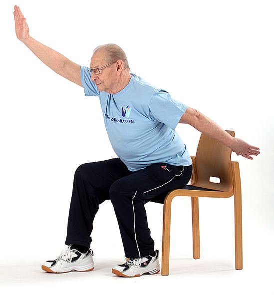 Tuolin reunalla istuva jumppaaja nojaa ylävartalolla eteenpäin ja on ojentanut vasemman käden suoraksi eteen yläviistoon ja oikean käden taakse alaviistoon.