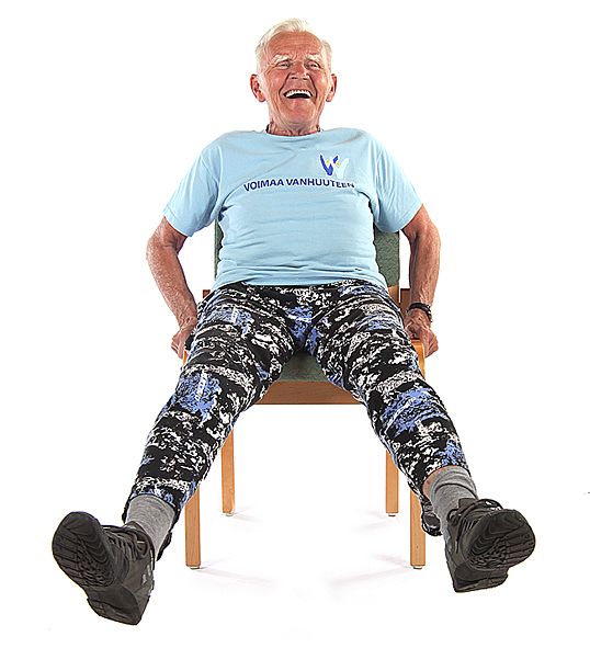 Jumppaaja istuu tuolilla jalat ojennettuina eteen ja vie jalkoja ilmassa sivulle pitäen samalla käsillä kiinni tuolin reunoista.