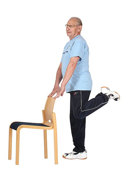 Jumppaaja seisoo ryhdikkäästi pitäen käsillä kiinni tuolin selkänojasta ja nostaa vasemman jalan kantapäätä kohti pakaraa