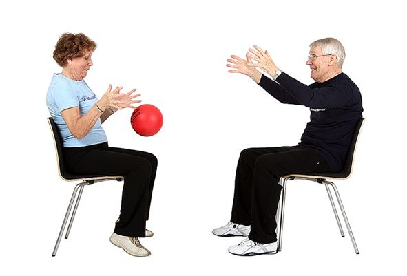Kaksi jumppaajaa istuu kasvotusten tuoleilla jalat pienessä haara-asennossa. Toinen jumppaaja on pompauttanut pallon lattian kautta toiselle jumppaajalle