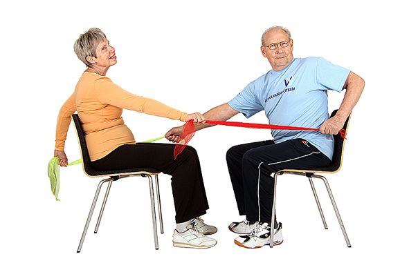 Kaksi jumppaajaa istuu tuoleilla kasvotusten ja pitää vastakkaisilla käsillä saman huivin päistä kiinni. Toinen jumppaaja on vetänyt oikean tai toinen vasemman kyynärpään koukkuun selän taakse