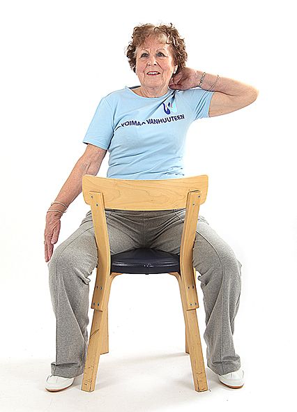 Jumppaaja istuu rintamasuunta tuolin selkänojaan päin ja on vienyt vasemman käden koukussa niskan taakse. Oikealla kädellä hän kurkottaa polven vierestä kohti lattiaa.