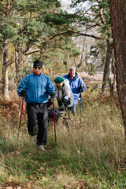 Muutama jumppaaja on kävelemässä jonossa metsässä polkua pitkin. Osa käyttää apunaan sauvoja tai rollaattoria.