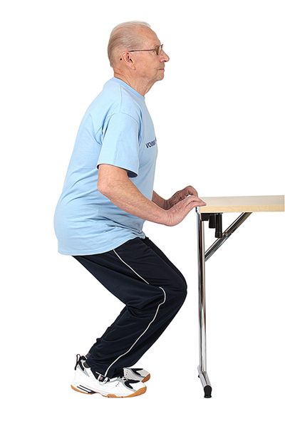 Jumppaaja on kyykistymässä ja ottaa sormenpäillä tukea vartalon edessä olevasta pöydänreunasta.
