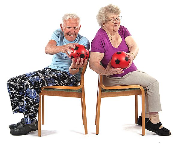 Kaksi jumppaajaa istuu tuoleilla selät vastakkain. Toinen jumppaajista kiertää ylävartaloa oikealla ja toinen vasemmalle yrittäen koskettaa pallolla toisen palloa.
