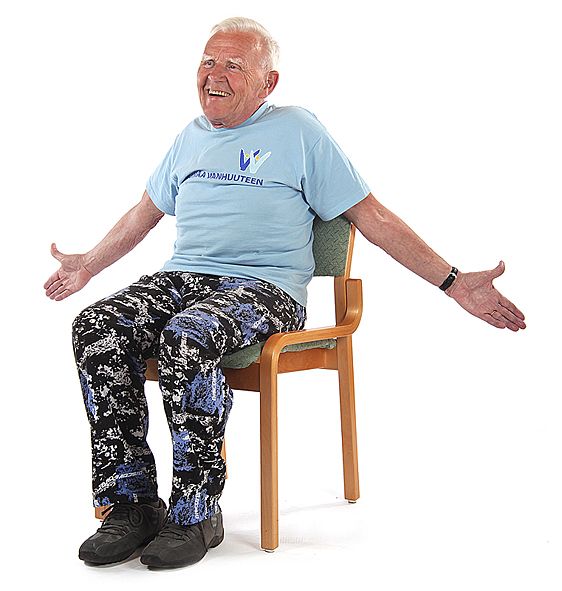 Jumppaaja istuu ryhdikkäästi tuolilla ja on avaamassa rintakehää vieden käsiä suorana sivukautta taaksepäin peukaloiden osoittaessa kohti kattoa.
