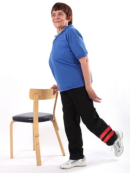 Jumppaaja seisoo ryhdikkäästi oikealla jalalla ja on ojentanut vasemman jalan suorana taakse pitäen samalla oikealla kädellä kiinni tuolin selkänojasta. Vasemmassa jalassa on nilkkapaino.