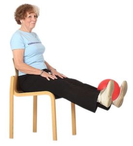 Jumppaaja istuu tuolilla pitäen käsiä reisien päällä ja on ojentanut molemmat jalat eteen suoriksi irti lattiasta pitäen palloa nilkkojen välissä.