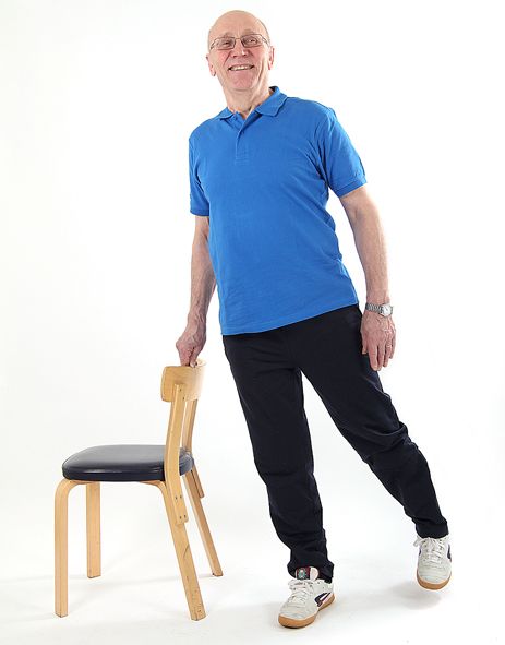 Jumppaaja seisoo tuolin vieressä oikealla jalalla ja on nostamassa vasenta jalkaa sivulle kantapää edellä ottaen samalla kevyesti tukea tuolin selkänojasta.