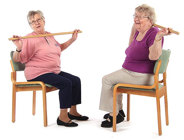 Kaksi jumppaajaa istuu tuoleilla vastakkain toisiaan ja toinen jumppaajista pitää käsissään olevaa puukeppiä rintakehällä ja toinen pitää omaansa niskan takana. He kiertävät ylävartaloa sivulle katseen seuratessa liikkeen suuntaan.
