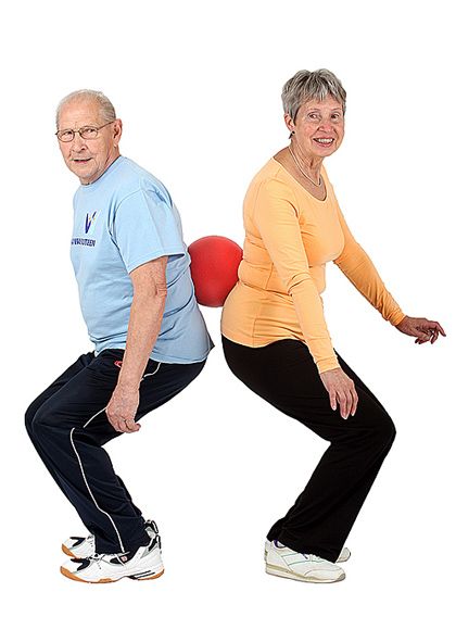 Kaksi jumppaajaa seisoo pienessä haara-asennossa selin toisiinsa pallon ollessa heidän välissään alaselän kohdalla ja ovat kyykistymässä yhtäaikaa nojaten samalla palloon.