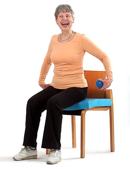 Jumppaaja istuu tuolilla olevan tasapainotyynyn päällä ja on antamassa vasemmalla kädellä palloa selän takaa oikeaan käteen.