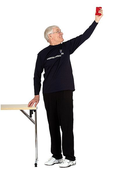 Jumppaaja seisoo pitäen oikealla kädellä kiinni vieressä olevasta pöydästä ja on ojentanut vasemman käsivarren suoraksi ylös takaviistoon. Hän on kääntänyt pään vasemmalle katsoakseen vasemmassa kädessä olevaa hernepussia.