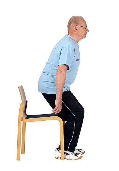 Jumppaaja on nousemassa tuolilta ylös ojentamalla jalkoja ja nojaamalla ylävartalolla eteenpäin katsoen samalla eteenpäin.