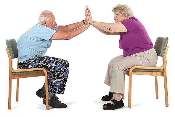Kaksi jumppaajaa istuu tuoleilla kasvotusten noin metrin päässä toisistaan. He ovat kumartaneet eteenpäin ja ojentaneet molemmat kätensä suoraksi eteen koskettaen molemmilla käsillä toistensa vastakkaisia käsiä.