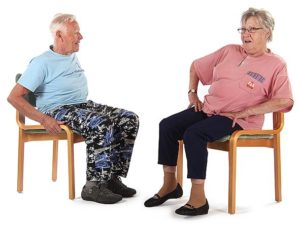 Kaksi jumppaaja istuu tuoleilla toisiaan vastakkain pitäen käsillä kiinni lanteista tai tuolin reunoista