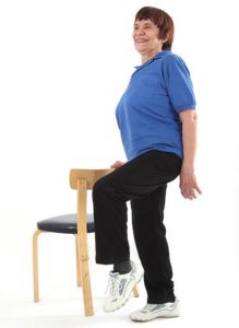 Jumppaaja seisoo vasemmalla jalalla ja on nostanut oikean jalan irti lattiasta nostamalla polvea ylöspäin. Hän ottaa oikealla kädellä tukea vartalon vieressä olevan tuolin selkänojasta ja katsoo eteenpäin.