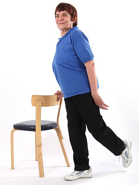 Jumppaaja seisoo ryhdikkäästi oikealla jalalla ja on ojentanut vasemman jalan suorana taakse pitäen samalla oikealla kädellä kiinni tuolin selkänojasta.