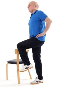 Jumppaaja seisoo ryhdikkäästi vasemmalla jalalla ja on nostanut oikean polven ylös ottaen samalla kädellä tukea vieressä olevan tuolin selkänojasta.