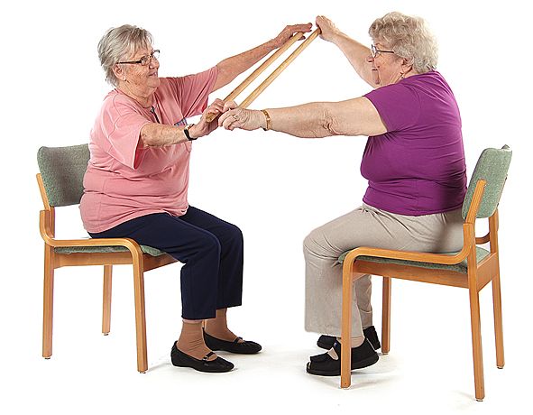 Kaksi jumppaajaa istuu tuoleilla toisiaan vastakkain ja pitävät kumpikin puukeppiä käsissään. Jumppaajat ovat nostamassa puukeppiä yhtäaikaa ylös käsien ollessa yläviistossa vartalon edessä.