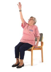 Jumppaaja istuu ryhdikkäästi tuolissa ja on ojentanut oikean käden suoraksi ylös katseen seuratessa kättä.