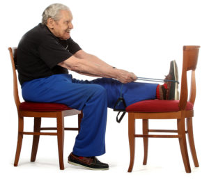 Jumppaaja istuu tuolilla ja on ojentanut vasemman jalan suoraksi edessä olevalle toiselle tuolille. Hän vetää ylävartaloa suorana olevaa jalkaa kohti käyttäen apunaan käsissään olevaa kuminauhaa