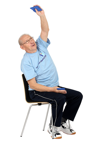 Jumppaaja istuu tuolilla pitäen molemmissa käsissä hernepussia ja on ojentanut vasemman käden suoraksi ylös
